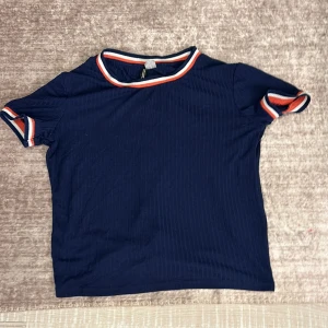 Mörkblå ribbad topp från H&M Divided - Säljer en mörkblå ribbad topp från H&M Divided i storlek M. Den har korta ärmar och en rund halsringning med ränder i vitt och orange. Perfekt för en avslappnad vardagslook! Gammal men ser oanvänd ut.