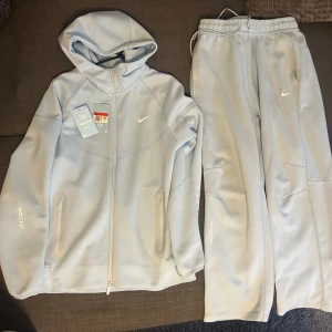 Ljusgrå hoodie och byxor från NOCTA - OBS passar mer som M än L! Men säljer ett set med ljusblå hoodie och matchande byxor från NOCTA. Hoodien har dragkedja och en stilren design med Nike-logga. Perfekt för en avslappnad stil eller träning. Kommer med taggar. Kan finnas nån liten fläck som kan vara svår att ta bort, men inget man tänker på. 