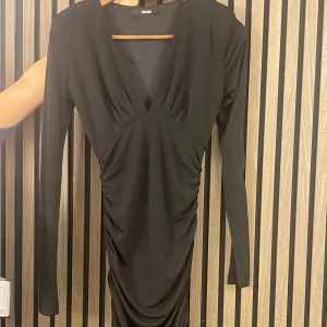 Svart långärmad klänning  - Snygg svart klänning från BikBok med lång ärm och v-ringad design. Perfekt för en kväll ute eller en fest. Den har en smickrande passform med rynkade detaljer som ger en elegant look. Passar bra till både höst och vinter. Har axelvaddar. Nypris: 600kr