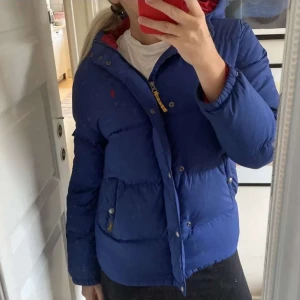 Blå dunjacka med huva - Säljer en blå dunjacka med huva, perfekt för kalla dagar. Jackan har en dragkedja och knappar framtill. Den är i bra skick men har ett litet hål på framsidan. Passar bra för höst och vinter.