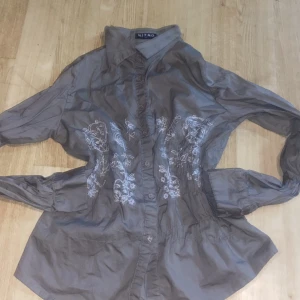 Grå skjorta från Mitno - Säljer en snygg grå skjorta från Mitno i storlek XL. Skjortan har långa ärmar och är dekorerad med fina broderade blommor på framsidan. Perfekt för både vardag och festliga tillfällen. Knappar framtill för en klassisk look. Passar bra till både jeans och kjol!