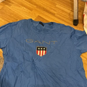 Blå t-shirt från GANT - Säljer en snygg blå t-shirt från GANT i storlek L. Den har ett tryck med GANT-loggan och ett sköldemblem med stjärnor och ränder på framsidan. Perfekt för en avslappnad stil. T-shirten är i bra skick och gjord av mjuk bomull.