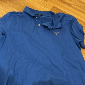 Blå pikétröja från GANT - Säljer en snygg blå pikétröja från GANT i storlek L. Tröjan har en klassisk krage med två knappar och GANT-loggan broderad på bröstet. Perfekt för både vardag och lite finare tillfällen. Passar bra till jeans eller chinos!