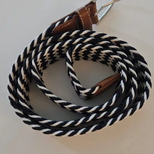 Flätat bälte i blått och vitt - Säljer ett snyggt flätat bälte i blått och vitt med bruna läderdetaljer. Perfekt för att ge din outfit en cool touch. Bältet har en robust känsla och passar både till vardags och fest. Spännet är i silverfärgad metall. Passar perfekt till jeans eller chinos!