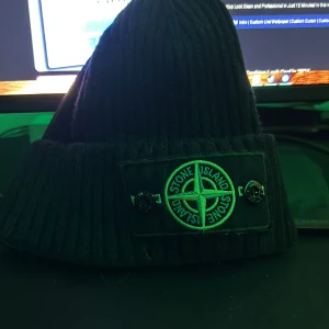 Svart mössa från Stone Island - Snygg svart mössa från Stone Island med deras ikoniska logga framtill. Perfekt för att hålla sig varm och stilren under kyliga dagar. Mössan är ribbad och har en klassisk design som passar till det mesta. Ett måste för alla som älskar streetwear! Säljer pga sugen på nåt nytt! Pris ej hugget i sten