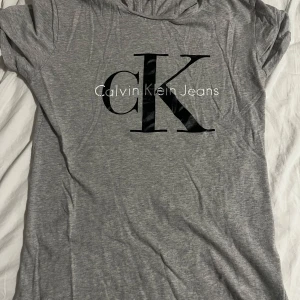 Grå t-shirt från Calvin Klein Jeans - Säljer en snygg grå t-shirt från Calvin Klein Jeans i storlek M. Den har en klassisk passform med korta ärmar och ett stort CK-logotryck på framsidan. Perfekt för en avslappnad stil eller som basplagg i garderoben. 🖤priset går att diskutera!