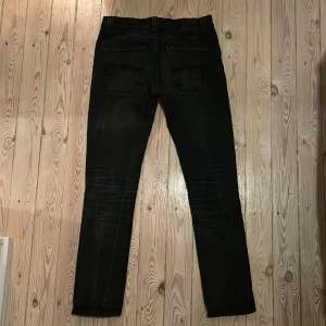 Nudie Jeans Grim Tim - Säljer ett par svarta jeans i modell Grim Tim från Nudie Jeans i mycket bra skick. De har en slim passform och är perfekta för både vardag och fest. Jeansen är tillverkade i bomull och har en klassisk design med fickor och knappgylf. Perfekta för höst och vinter!