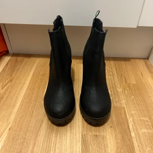 Svarta boots med klack - Snygga svarta boots i syntetmaterial med hög klack och grov sula. Perfekta för höst och vinter, dessa boots har en stilren design utan snörning eller dragkedja. De är helt nya aldrig använda kolla bild på sulan och passar perfekt till både vardag och fest. Enkla att matcha med olika outfits! Storlek 39. Skickas med spårbar frakt men går även att hämta i Malmö. 