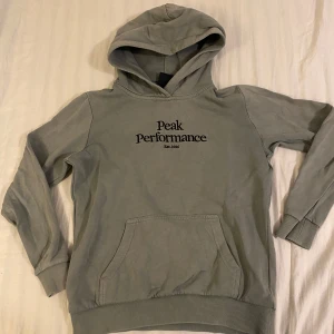 Grå/grön hoodie från Peak Performance - Säljer en snygg grå hoodie från Peak Performance i storlek 150. Den har en klassisk design med logga på bröstet och en praktisk magficka. Perfekt för både skola och fritid. Huvan är mjuk och bekväm, och tröjan är i bra skick. Passar perfekt till höst och vår!