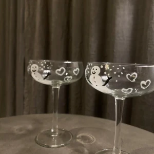 Cocktailglas med snögubbar  - Två eleganta champagneglas med charmigt vintertema. Glasen har söta snögubbar och hjärtan målade i vitt, vilket ger en festlig känsla. Perfekta för vinterfester eller som en söt detalj i hemmet. 85kr/styck eller 2 för 150kr