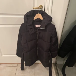 Svart pufferjacka från Nelly - Säljer en svart pufferjacka från Nelly i mycket bra skick. Jackan har en dragkedja framtill och ett bälte i midjan för en snygg passform. Jackan är knappt använd och är i mycket bra skick😊jackan är i XS men funkar för S