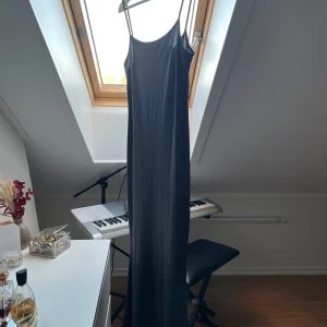 Grå långklänning med smala axelband - Elegant grå långklänning med smala axelband, perfekt för festliga tillfällen. Klänningen har en enkel och stilren design som passar både till vardags och fest. Materialet är mjukt och följsamt, vilket ger en bekväm passform.