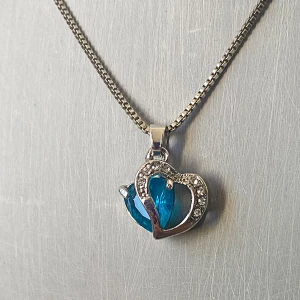 Halsband med blått hjärthänge - Snyggt halsband med ett hjärthänge i blått och silver. Hänget har en glittrig kant som ger en extra touch av elegans. Perfekt för att piffa upp vilken outfit som helst, oavsett om det är till vardags eller fest. Kedjan är i silverfärg och har en enkel låsning. Okänd metall 