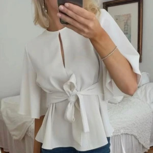 Vit blus med knytband - Snygg vit blus med knytband i midjan och korta ärmar. Perfekt för både vardag och fest. Den har en stilren design med en liten öppning framtill som ger en elegant touch.