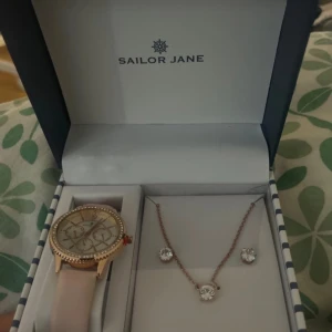 Smyckeset med klocka och halsband från Sailor Jane - Elegant smyckeset från Sailor Jane med en stilren klocka i guld och ett matchande halsband med örhängen. Klockan har en ljus urtavla och ett guldarmband, medan halsbandet och örhängena har glittrande stenar. Perfekt för både vardag och fest!