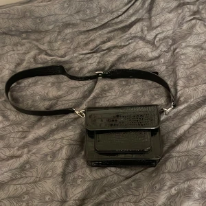 Svart axelväska i krokodilmönster - Snygg svart axelväska med krokodilmönster. Väskan har en justerbar axelrem och flera fack för enkel organisering. Perfekt för både vardag och fest. Den glänsande ytan ger en lyxig känsla. Passar till alla säsonger och outfits!