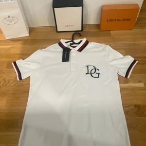 Vit pikétröja från Dolce & Gabbana - Helt ny !! Säljer en snygg vit pikétröja från Dolce & Gabbana. Den har en broderad logga på bröstet och stiliga ränder i svart och rött på krage och ärmslut. Perfekt för en casual men ändå uppklädd look. Tröjan är i bomull och känns riktigt lyxig. Passar perfekt till både vardag och fest!