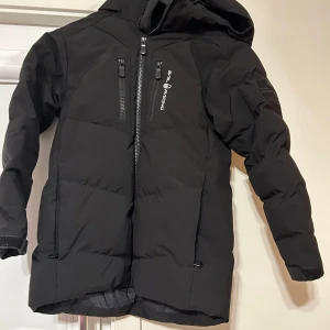 Svart dunjacka från Sail Racing - Säljer en svart dunjacka från Sail Racing i mycket bra skick. Jackan har en stilren design med dragkedja och flera praktiska fickor. Perfekt för kalla vinterdagar med sin varma och skyddande huva. Passar både till vardags och utomhusaktiviteter.