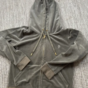Grå hoodie från Juicy Couture - Säljer en supermjuk och snygg grön hoodie från Juicy Couture i storlek S. Den har en dragkedja med ett coolt J-hänge och justerbara snören i luvan. Perfekt för en avslappnad stil eller mysiga dagar hemma. 🩶