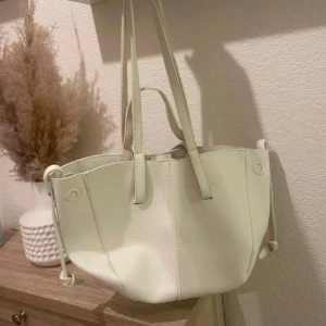 Vit axelväska i skinn - Snygg vit axelväska i skinn med stilren design. Väskan har två långa handtag och en praktisk storlek för vardagsbruk. Perfekt för att bära med sig det nödvändigaste med stil.