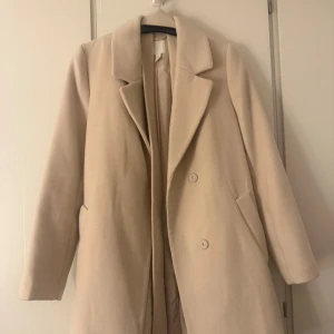 Beige kappa från H&M - Säljer en stilren beige kappa från H&M i storlek M. Den har en klassisk design med knappar framtill och långa ärmar. Perfekt för höst och vår, och ger en elegant touch till vilken outfit som helst. Kappan är i mycket bra skick och redo för nya äventyr!