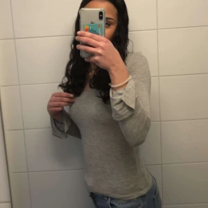 Grå långärmad topp - Säljer en stilren grå långärmad topp i mjukt material. Perfekt för vardagsbruk eller att klä upp med accessoarer. Toppen har en enkel design med en bekväm passform. Passar bra till jeans eller kjol för en avslappnad look. Köpt från gina och är i bra skick.