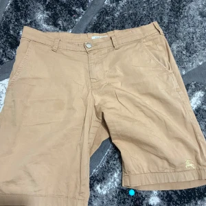 Beige shorts från Burberry - Säljer ett par snygga beige shorts från Burberry. De har en klassisk design med dragkedja och knapp framtill. Perfekta för sommaren med sin stilrena look och bekväma passform. Märkesloggan är diskret broderad på baksidan. Passar både till vardags och lite finare tillfällen.