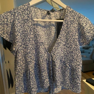Blå blommig blus - Söt blå blommig blus med korta ärmar och v-ringning. Den har en knytning framtill som ger en fin detalj 🌸