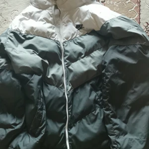 Grå och vit pufferjacka från Nike - Säljer en snygg och varm pufferjacka från Nike i grått och vitt. Jackan har en dragkedja framtill och är perfekt för kyliga dagar. Den är i mycket bra skick och passar perfekt för både höst och vinter. Huvtröjan ger extra skydd mot väder och vind. Perfekt för dig som vill hålla stilen även när det är kallt ute!