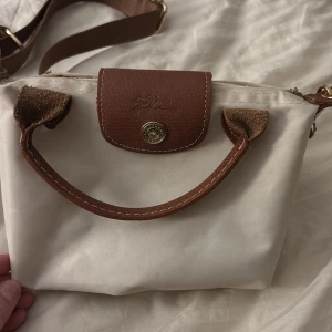 Beige och brun axelväska - Säljer en snygg vit axelväska med bruna detaljer. Väskan har en praktisk dragkedja och ett elegant brunt lock med knapp. Perfekt för vardagsbruk eller en dag på stan. Den är i mycket bra skick och redo för nya äventyr!