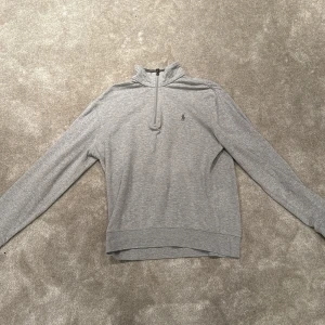 Grå tröja från Ralph Lauren - Säljer en stilren grå tröja från Ralph Lauren i mycket bra skick. Den har en dragkedja vid halsen och det klassiska Ralph Lauren-logotypen broderad på bröstet. Perfekt för både vardag och lite finare tillfällen. Passar bra till höst och vår!
