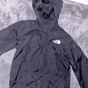 Svart vindjacka från The North Face - Säljer en svart vindjacka från The North Face i mycket bra skick. Jackan har en praktisk huva och dragkedja framtill. Perfekt för blåsiga dagar och passar bra till både vår och höst. Den är långärmad och gjord i ett lätt syntetmaterial. Funkar väldigt bra i regnet och är i storlek XS