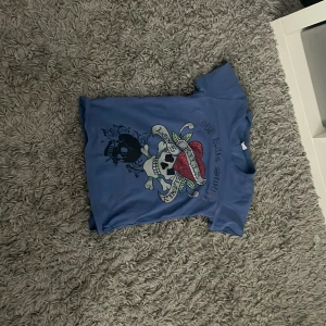 Blå t-shirt med tryck från Ed Hardy - Säljer en blå t-shirt från Ed Hardy med coolt tryck av en dödskalle med hatt och bandana på framsidan och en stor ros på baksidan. T-shirten är kortärmad och gjord av 100% bomull. Perfekt för dig som gillar unika och uttrycksfulla plagg!