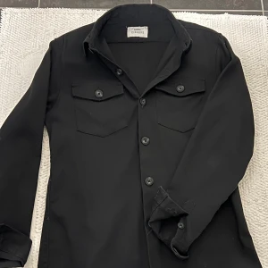 Svart overshirt från Ciszere - Snygg svart skjorta från Ciszere med långa ärmar och knappar framtill. Skjortan har två bröstfickor med knappar och en klassisk krage. 