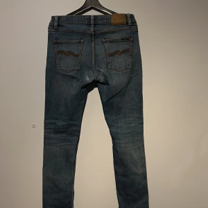 Blå jeans ifrån nudie med broderade bakfickor - Snygga blå jeans med broderade detaljer på bakfickorna. De har en klassisk femficksdesign och en knappgylf. Perfekta för en avslappnad stil. Passar runt 160-172