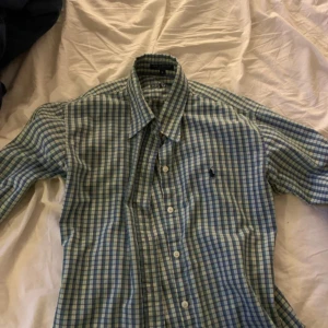Rutig skjorta från Ralph Lauren - Snygg rutig skjorta från Ralph Lauren i gröna och blå nyanser. Skjortan har långa ärmar och knappar framtill. Perfekt för en stilren look.