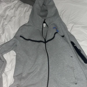 Grå hoodie från Nike med Barcelona-logga - Säljer en grå nike tech fleec med Barcelona-logga och texten 'CULERS' i blått och rött. Tröjan har en dragkedja framtill och en huva. Perfekt för fotbollsfans!