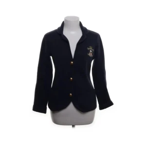 Mörkblå kavaj från Lexington - Säljer en stilren mörkblå kavaj från Lexington med tre guldknappar framtill och ett broderat emblem på bröstet. Kavajen har en klassisk krage och långa ärmar, perfekt för en elegant look.