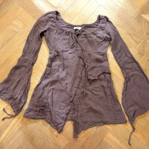 Asymmetrisk blus - Asymmetrisk supersöt lavendelfärgad blus i linne med snörning i ärmar och krage och blomma på ena sidan. 