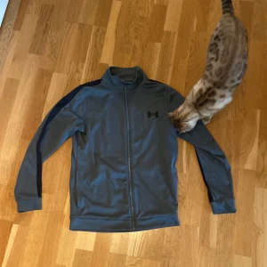 Grå träningsjacka från Under Armour - Säljer en grå träningsjacka från Under Armour med dragkedja och långa ärmar. Jackan har en stilren design med mörkare detaljer längs ärmarna och logotypen på bröstet. Perfekt för träning eller en aktiv vardag.