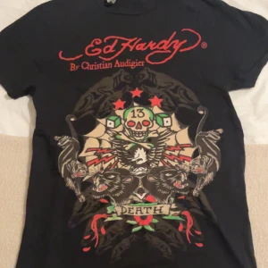 Svart t-shirt från Ed Hardy - Snygg svart t-shirt från Ed Hardy med ett stort tryck på framsidan som inkluderar dödskalle och texten 'Death'. Röd Ed Hardy-logga både fram och bak. Perfekt för en edgy stil.