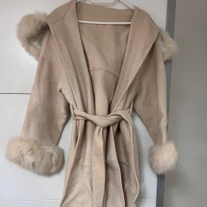 Beige kappa med pälsdetaljer  - Säljer en elegant beige kappa med äkta päls på huva och ärmslut. Kappan har en knytning i midjan och är perfekt för kyligare dagar. Den har en mjuk och lyxig känsla, vilket gör den till ett stilrent val för höst och vinter. S/M