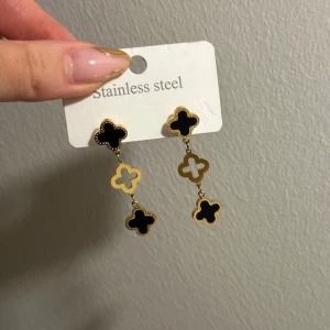 Svart och guld örhängen i rostfritt stål - Eleganta örhängen i rostfritt stål med svart och guld detaljer. Designen har en klöverformad stil med två hängande delar som ger en stilren look.