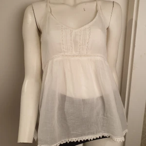 Vit ärmlös topp med spetsdetaljer - Säljer en söt vit ärmlös topp med tunna axelband och spetsdetaljer. Toppen har en luftig passform och dekorativa spetskanter längst ner. Insydd på sidorna, passar xs-s