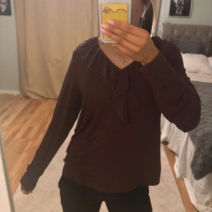 Volang blus - Säljer en elegant mörkbrun/vinröd långärmad topp med volang vid halsen. Perfekt för en stilren look. Toppen har en mjuk och bekväm passform.