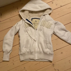 Vit hoodie från Abercrombie & Fitch - Säljer en vit hoodie från Abercrombie & Fitch med dragkedja och broderad logga på framsidan. Den har en mysig fodrad insida och ribbade muddar vid ärmslut och nederkant. Perfekt för kyliga dagar! Använt men fint skick, tvättas såklart vid köp!