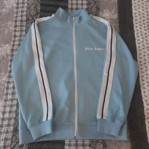 Ljusblå track jacket från Palm Angels - Säljer en snygg ljusblå track jacket från Palm Angels med vita och svarta ränder längs ärmarna. Jackan har en dragkedja framtill och en hög krage. Perfekt för en sportig look!