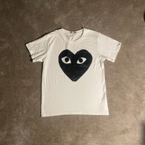 Vit t-shirt från Comme des Garçons - Säljer en vit t-shirt från Comme des Garçons med ett stort svart hjärta med ögon på framsidan. T-shirten är kortärmad och gjord av bomull. Unik och kommer göra dig stilren till sommaren.