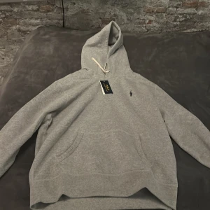 Grå hoodie från Ralph Lauren - Säljer en stilren grå hoodie från Ralph Lauren med klassisk logga på bröstet. Tröjan har en bekväm passform med känguruficka och justerbar huva. Perfekt för en avslappnad look. Nypris 2200 