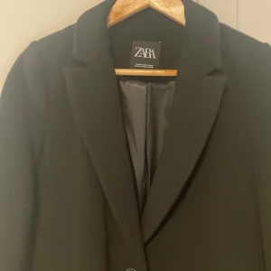 Svart kappa från Zara - Elegant svart kappa från Zara med klassisk design. Den har en enkel knäppning framtill och långa ärmar. Perfekt för en stilren look under kyligare dagar.